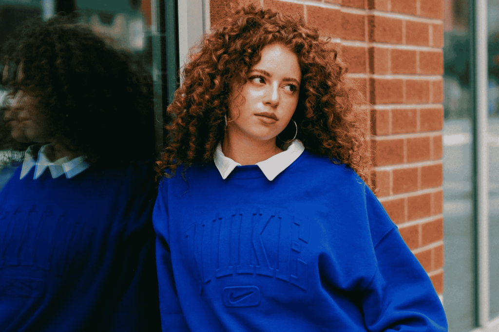 Woman with curly red hair in a blue Nike sweatshirt over a white collared shirt, leaning against a brick wall