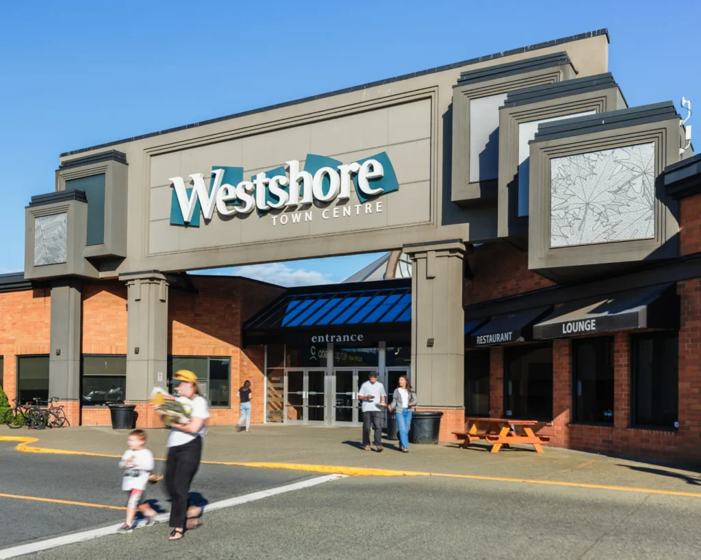 The main entrance of Westshore Town Centre under its sign on a sunny day, with shoppers walking out and a child crossing the parking lot